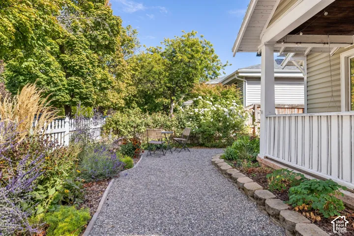Charming side yard pathway featuring gravel hardscaping, mature landscaping with lush greenery, flowering perennial beds, white picket fencing, and covered porch entry with white trim detailing
