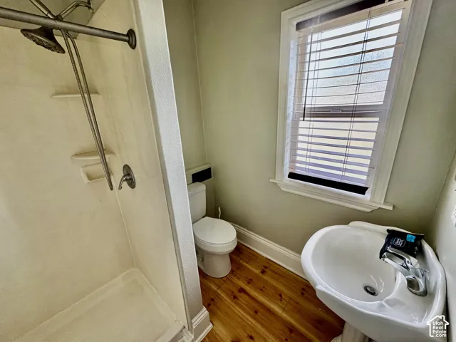 Walk-in shower with a chrome showerhead, a pedestal sink with a chrome faucet, toilet, wood-finish flooring, and a window with blinds