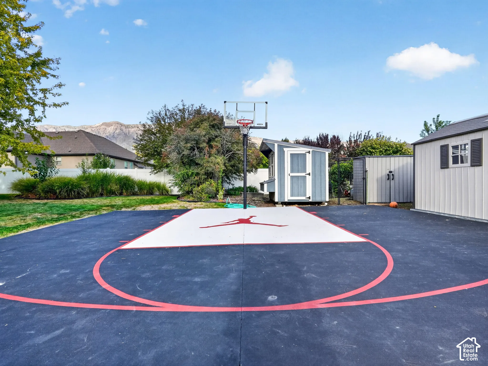 Hardscaped sports court with a basketball hoop, bordered by a white privacy fence and a manicured lawn