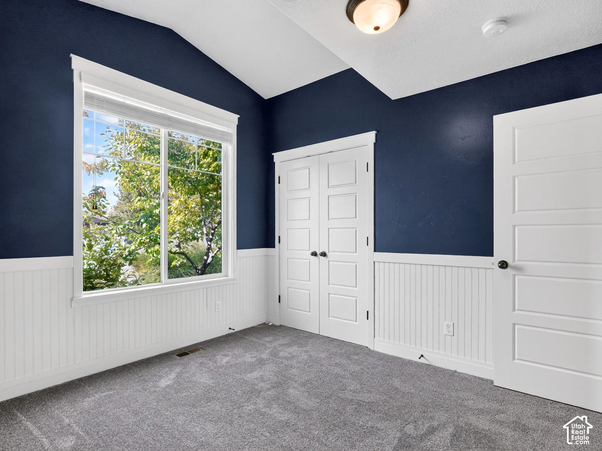 Vaulted ceiling with a flush-mount light fixture, large window, white wainscoting, gray carpet, and paneled doors