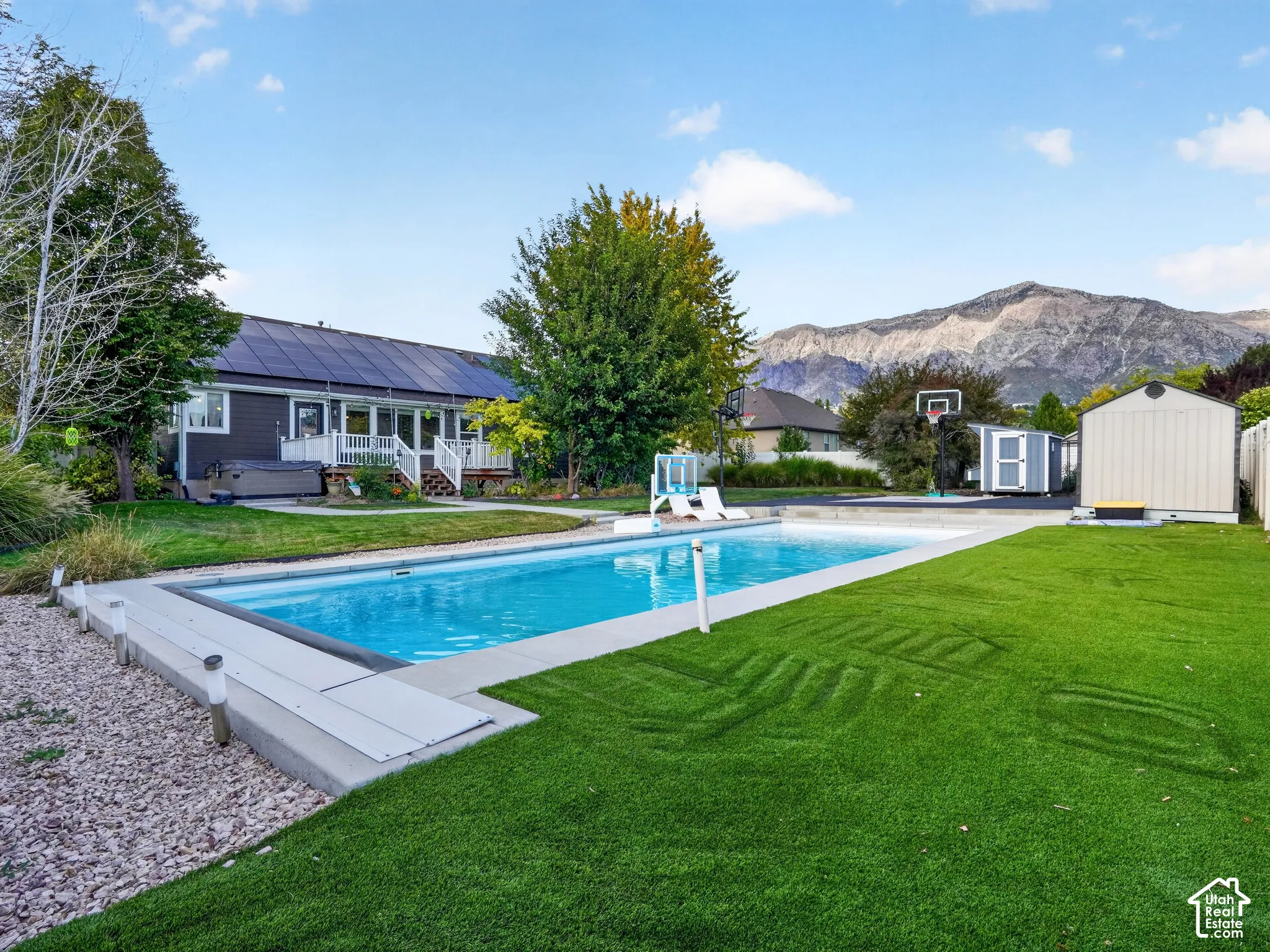 Expansive backyard featuring a rectangular swimming pool, artificial turf, two storage sheds, and a paved sport court