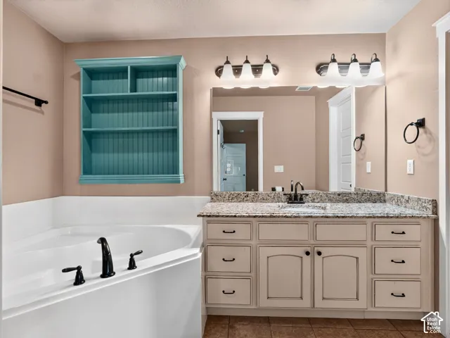 Built-in corner tub with dark fixtures, dual vanity with granite countertop, large mirror with vanity lighting, and wall-mounted shelving