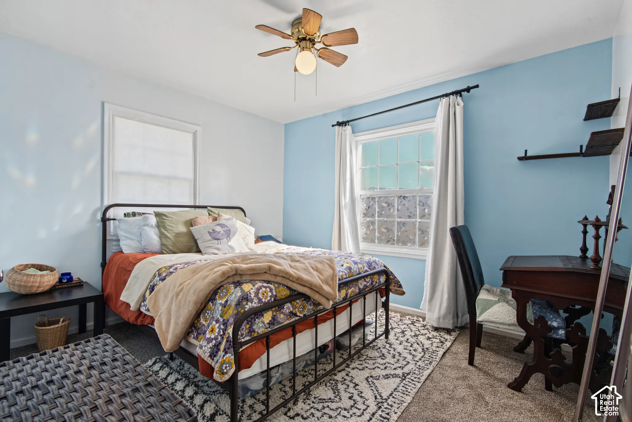 Bright secondary bedroom featuring soft blue accent walls, a ceiling fan with integrated lighting, large windows with natural light, patterned area rug, and black metal bed frame