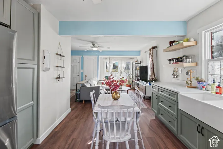 Bright, open-concept kitchen and dining area featuring wood-finish flooring, soft blue accent wall, ceiling fan, white shaker cabinetry with green painted lower cabinets, white countertops, stainless steel appliances, open shelving, and abundant natural l