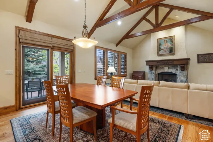Vaulted ceiling with exposed wood beams, stone fireplace, wood-finish flooring, sliding glass door access to an exterior deck, and a window with plantation shutters