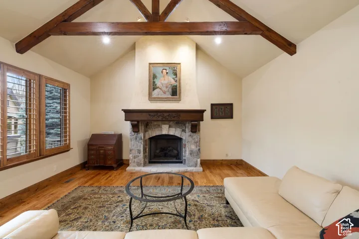 Vaulted ceiling with exposed wood beams, stone-clad fireplace with wood mantel, hardwood flooring, and plantation shutters