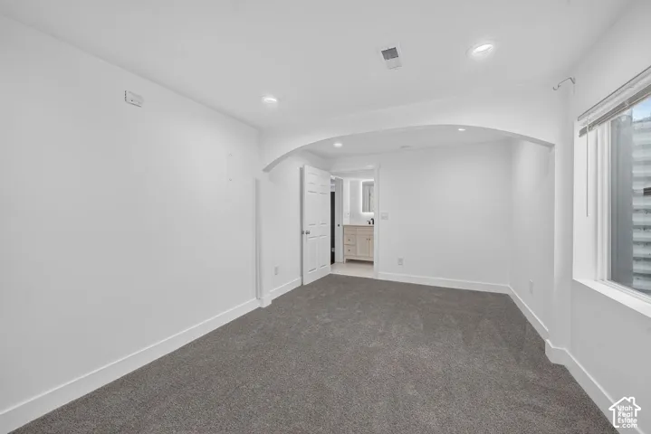 Spacious room featuring plush gray carpeting, recessed ceiling lighting, a window with blinds, and an arched entryway leading to a bathroom with a vanity