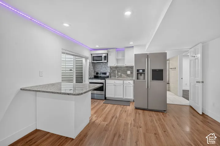 Kitchen featuring wood-finish flooring, stainless steel appliances, white cabinetry, a tile backsplash, and a breakfast bar with a granite-style countertop