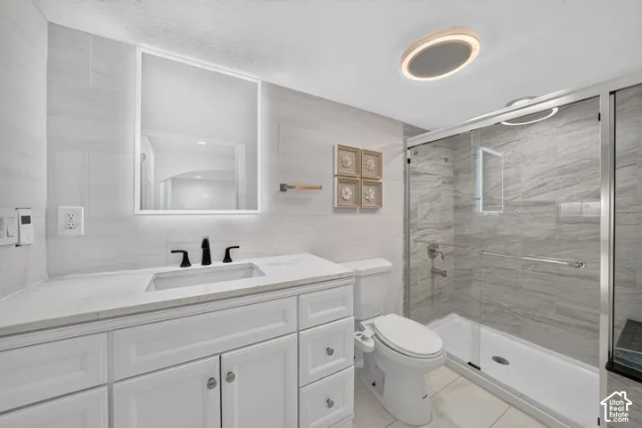 White vanity with integrated sink, modern matte black faucet, illuminated mirror, tiled shower with glass enclosure, and ceiling-mounted light fixture