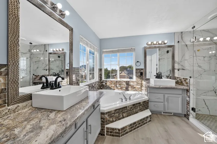Dual vanity with vessel sinks, built-in soaking tub, glass-enclosed shower, wood-finish flooring, and multiple windows
