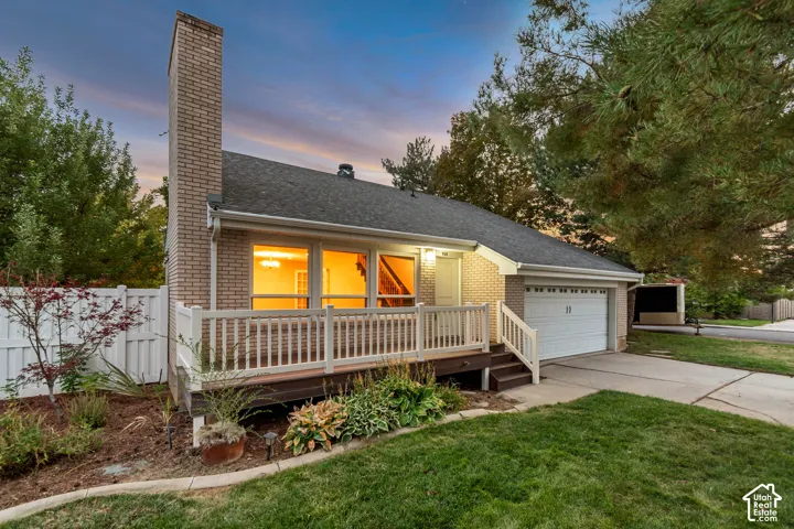 Brick exterior with a prominent chimney, front deck with white railing, attached garage, and a concrete driveway