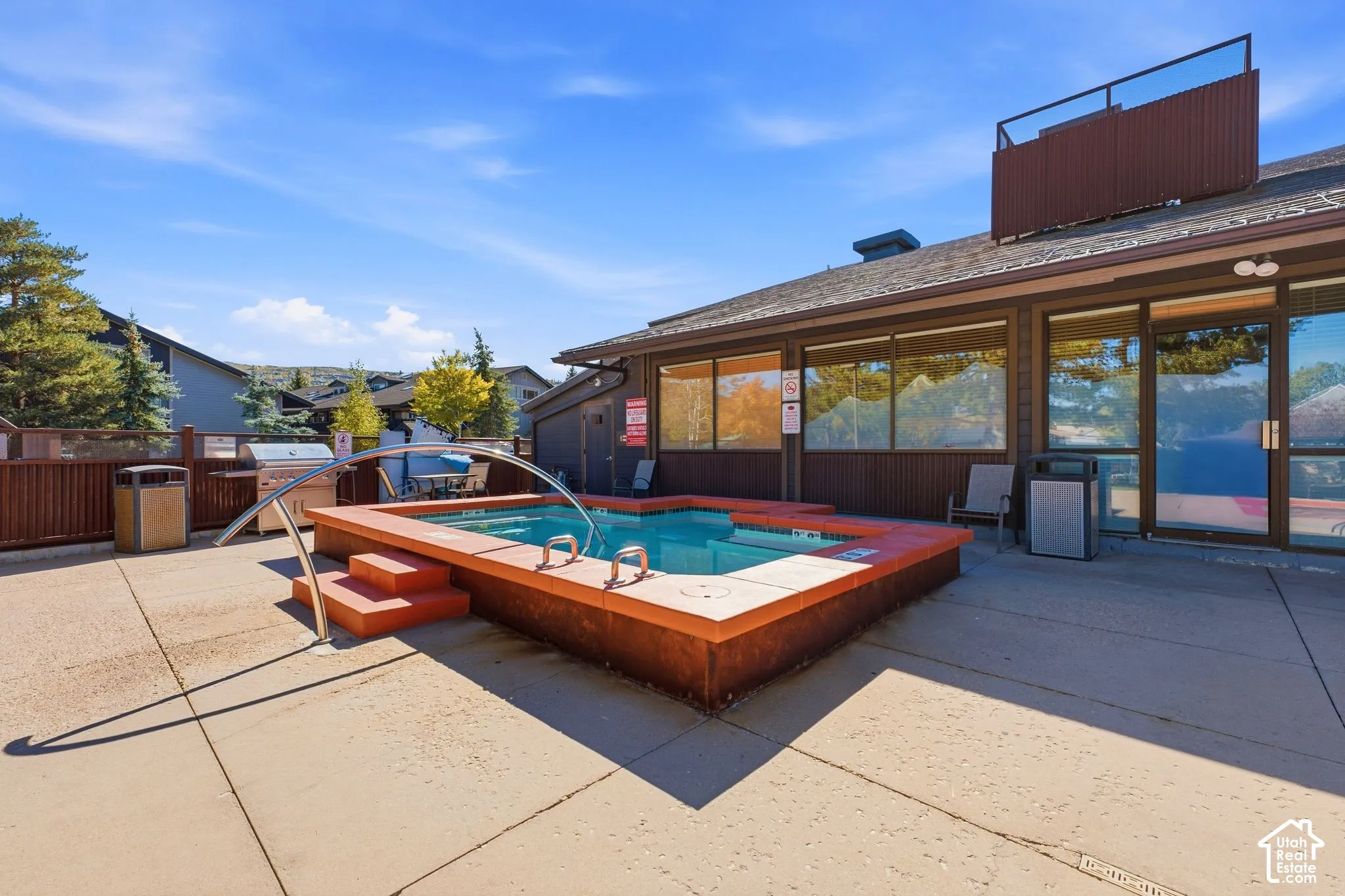 Outdoor hot tub with integrated steps and handrail, adjacent to a building featuring large windows and double glass doors