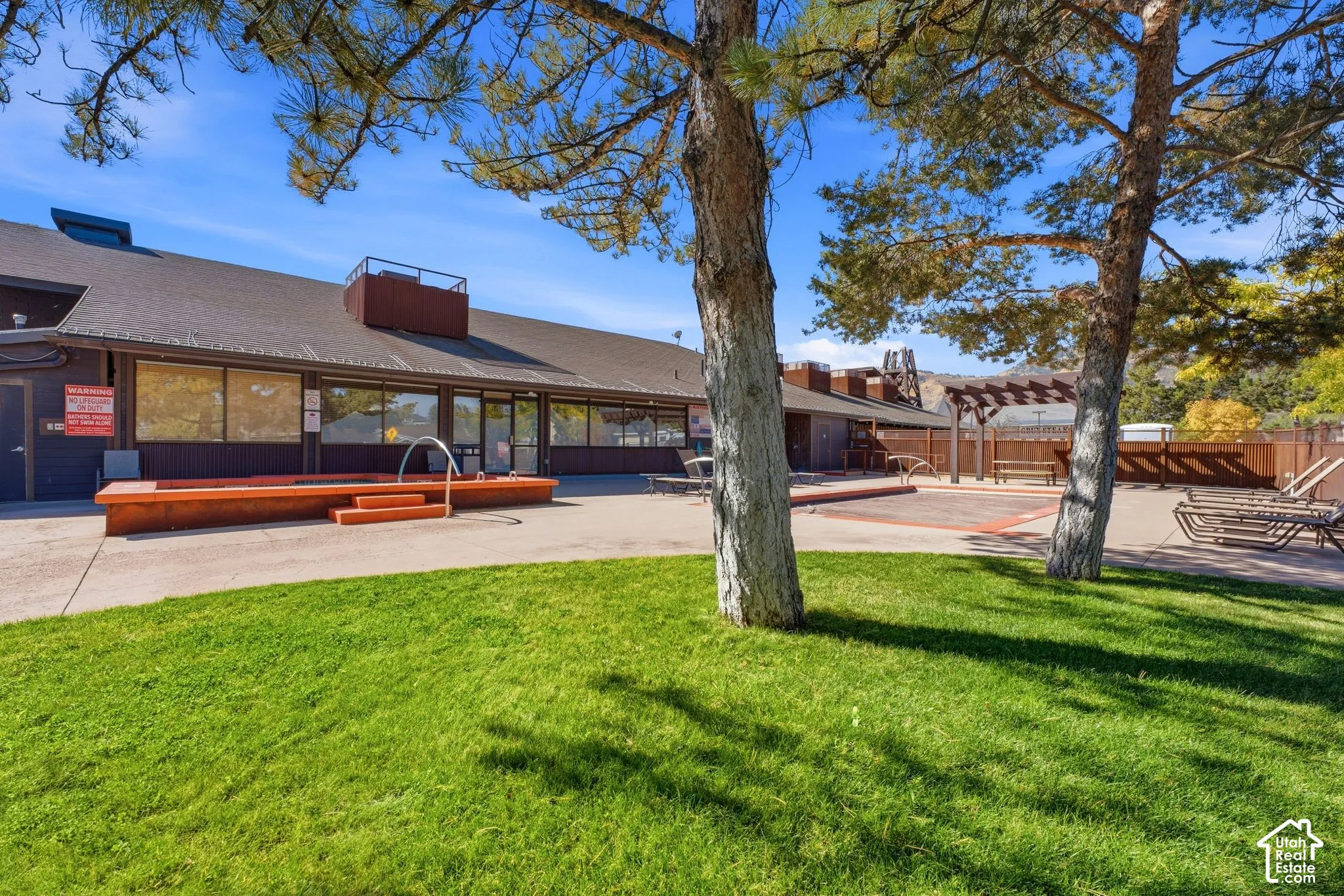 Manicured lawn area with mature trees, building with large windows, and an outdoor recreational space featuring a wooden pergola
