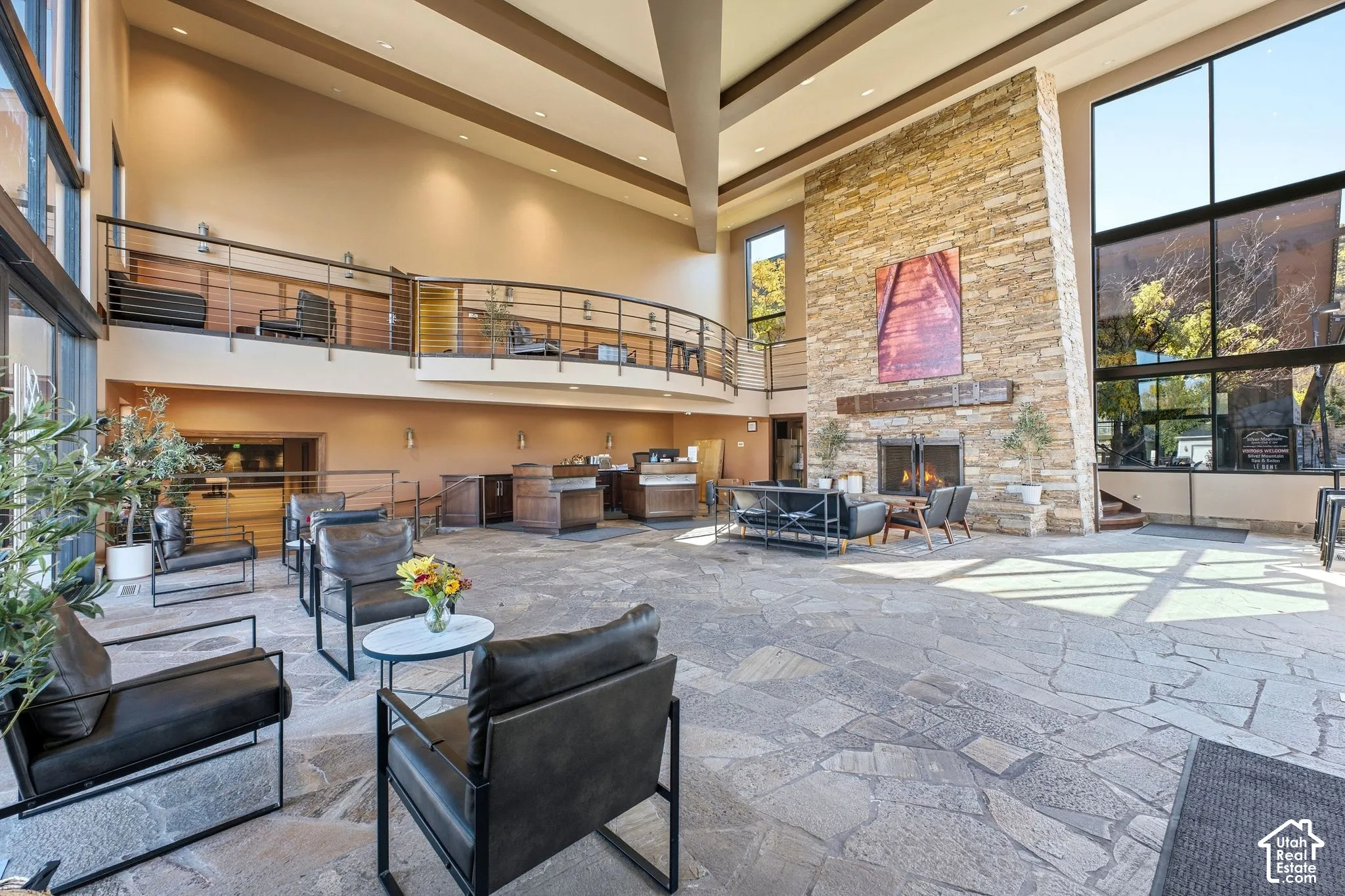 Vaulted ceilings with exposed beams, stone-clad fireplace, full-height windows, natural stone flooring, and an upper-level balcony with metal railing