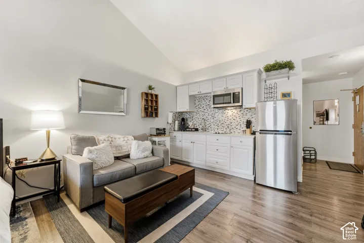 Vaulted ceiling above a compact living space featuring wood-finish flooring, white cabinetry, a mosaic tile backsplash, and stainless steel appliances