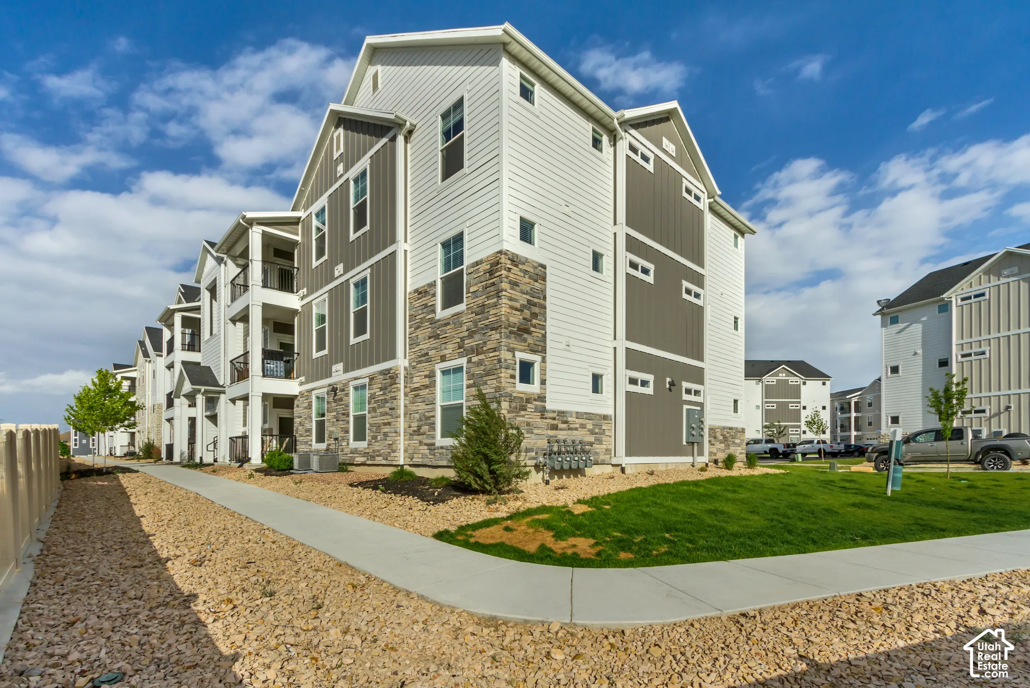 Multi-story building featuring a blend of horizontal siding and vertical paneling, stone veneer accents, individual balconies with dark railings, and a concrete pathway