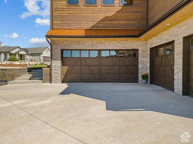 Wood-finish garage doors with window panels, stone veneer exterior, wood siding, concrete driveway, and stone retaining wall with steps