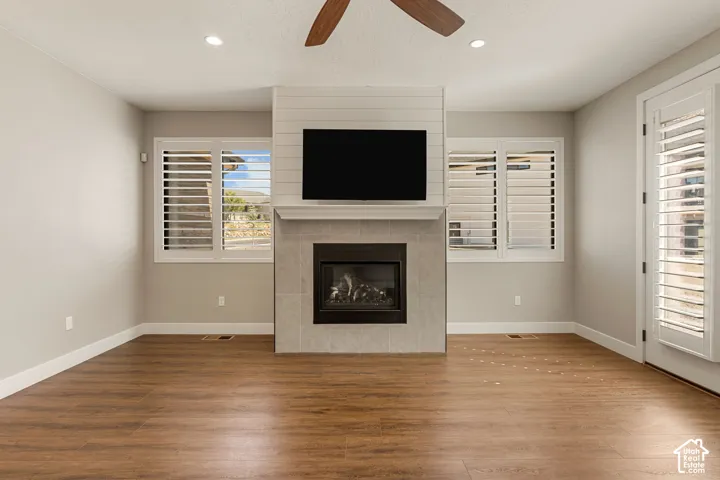 Spacious living room featuring a wood-finish plank floor, gas fireplace with stone surround and hearth, wall-mounted flat-screen television, ceiling fan, recessed lighting, and dual sets of white plantation shutters flanking the fireplace