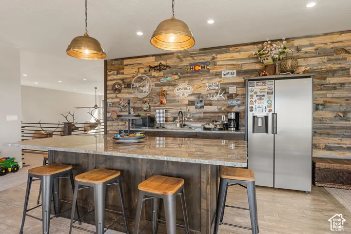 Expansive kitchen island with a stone-finish countertop, custom wood-paneled wall, stainless steel side-by-side refrigerator, recessed lighting, and industrial-style pendant lighting