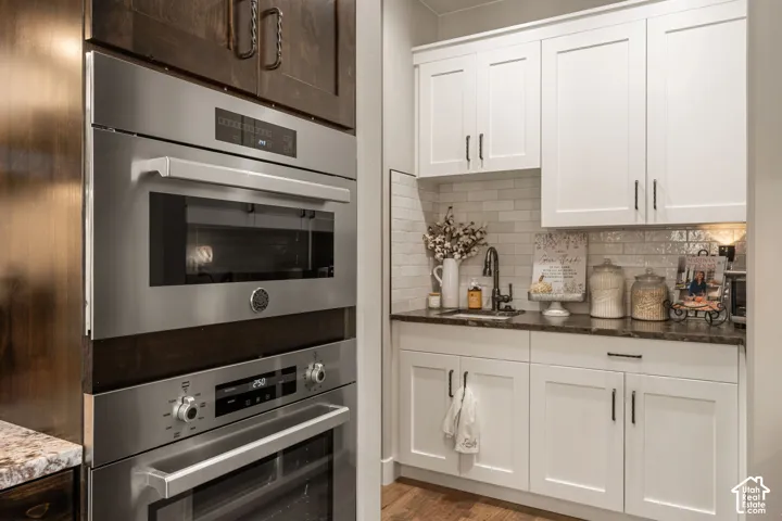 Built-in stainless steel double ovens, white shaker cabinetry, dark countertops, a subway tile backsplash, and a stainless steel sink with a dark faucet