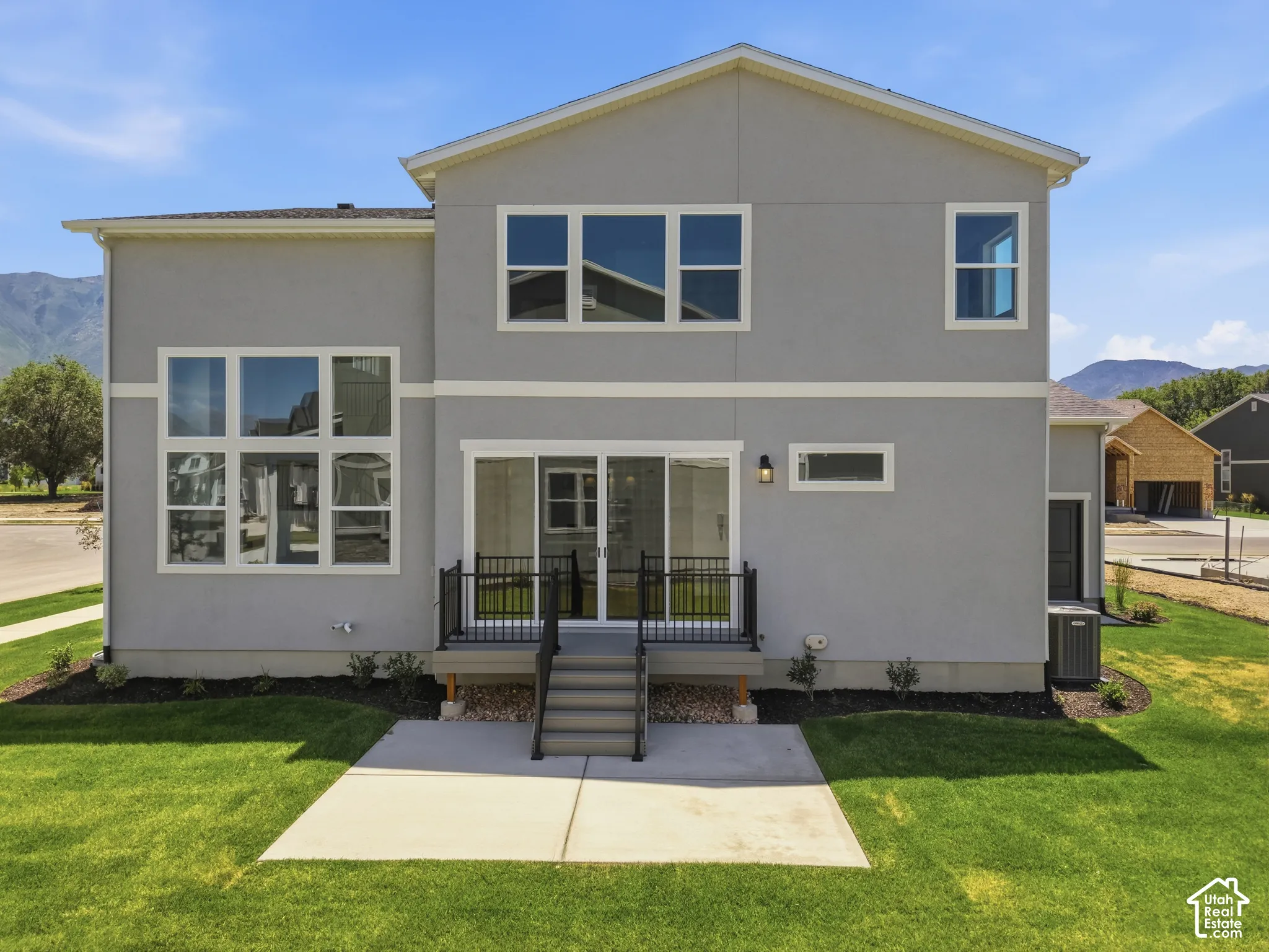 Contemporary stucco exterior with multiple large pane windows, a ground-level concrete patio, a raised deck with dark railing, and a sliding glass door entry