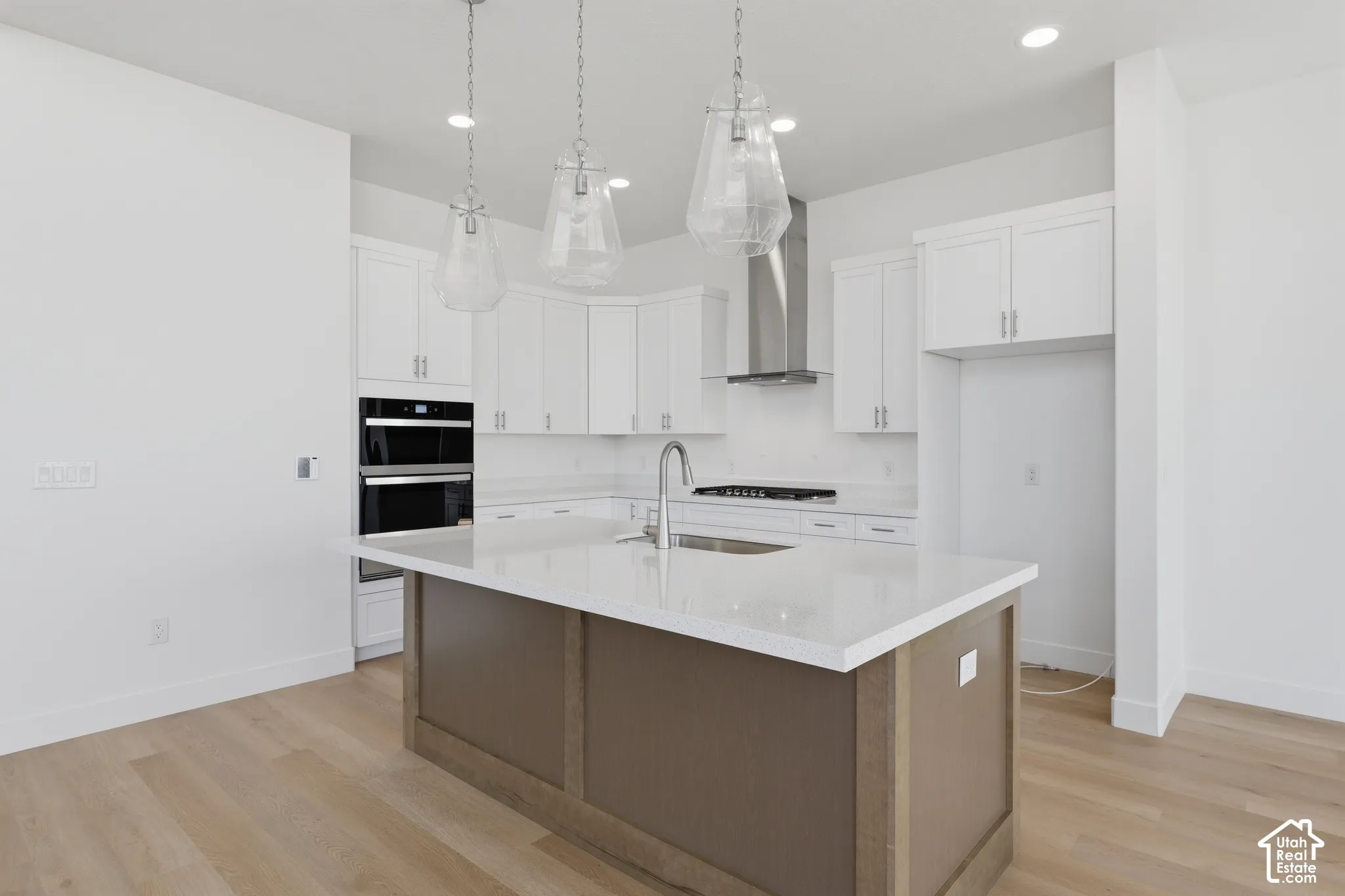 Open-concept kitchen featuring an expansive island with a light-toned countertop, white cabinetry, integrated stainless steel double ovens, a stainless steel range hood, and wood-finish flooring