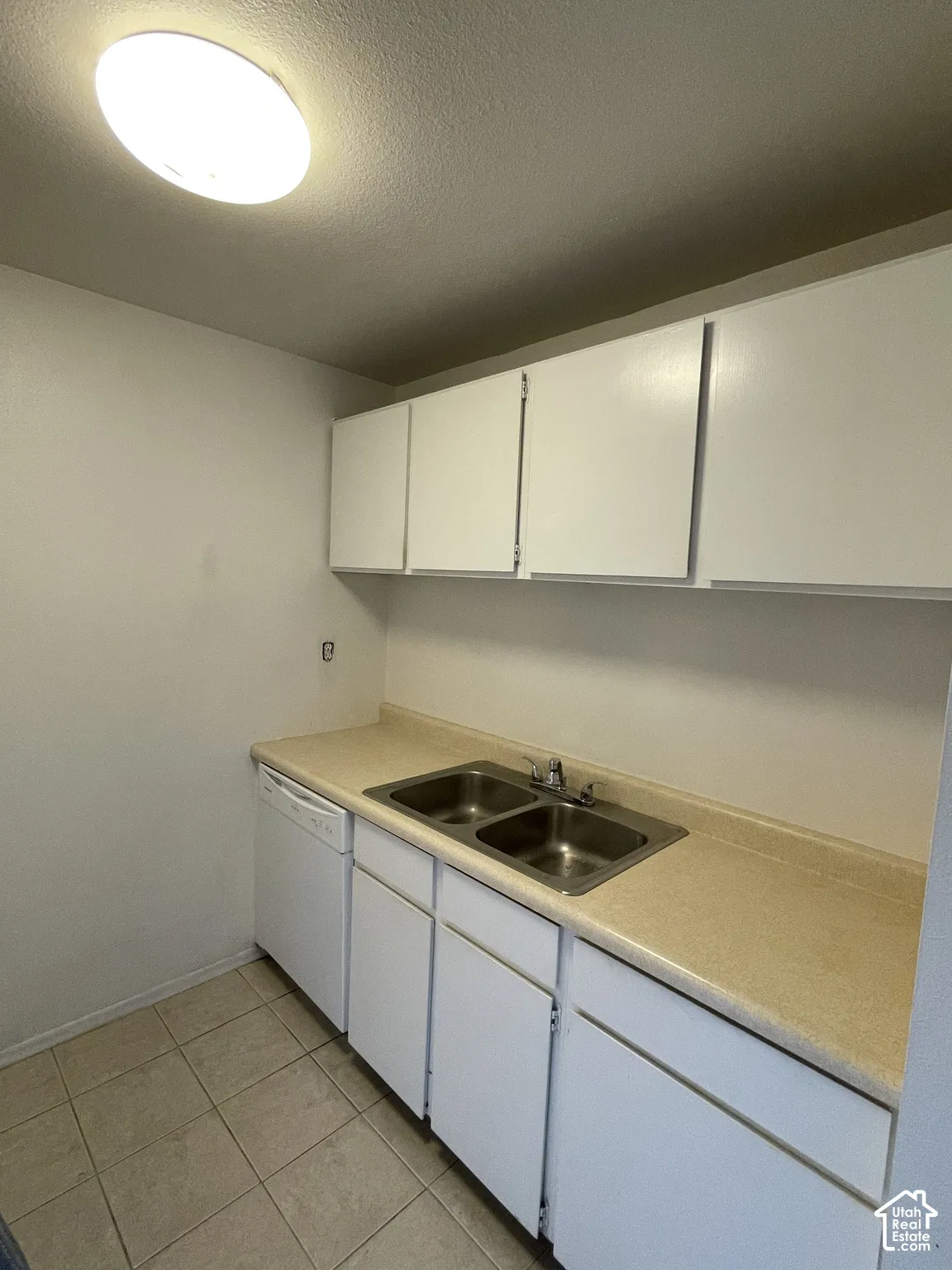 White cabinetry, dual basin stainless steel sink, light-toned countertop, white dishwasher, and tiled flooring