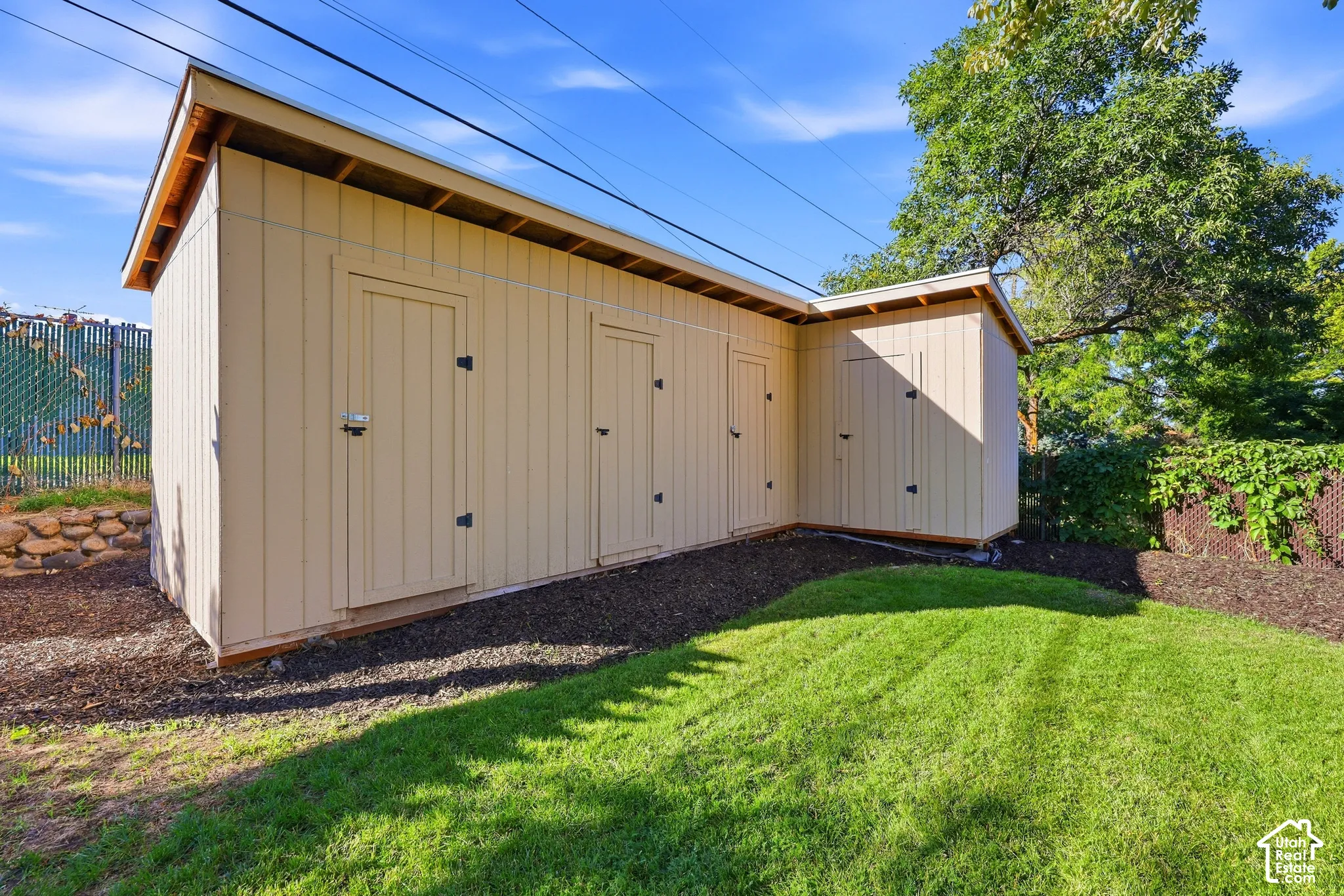 L-shaped shed with multiple access doors, a single-slope roof, and light-colored siding, situated on a sloped landscape with a manicured lawn