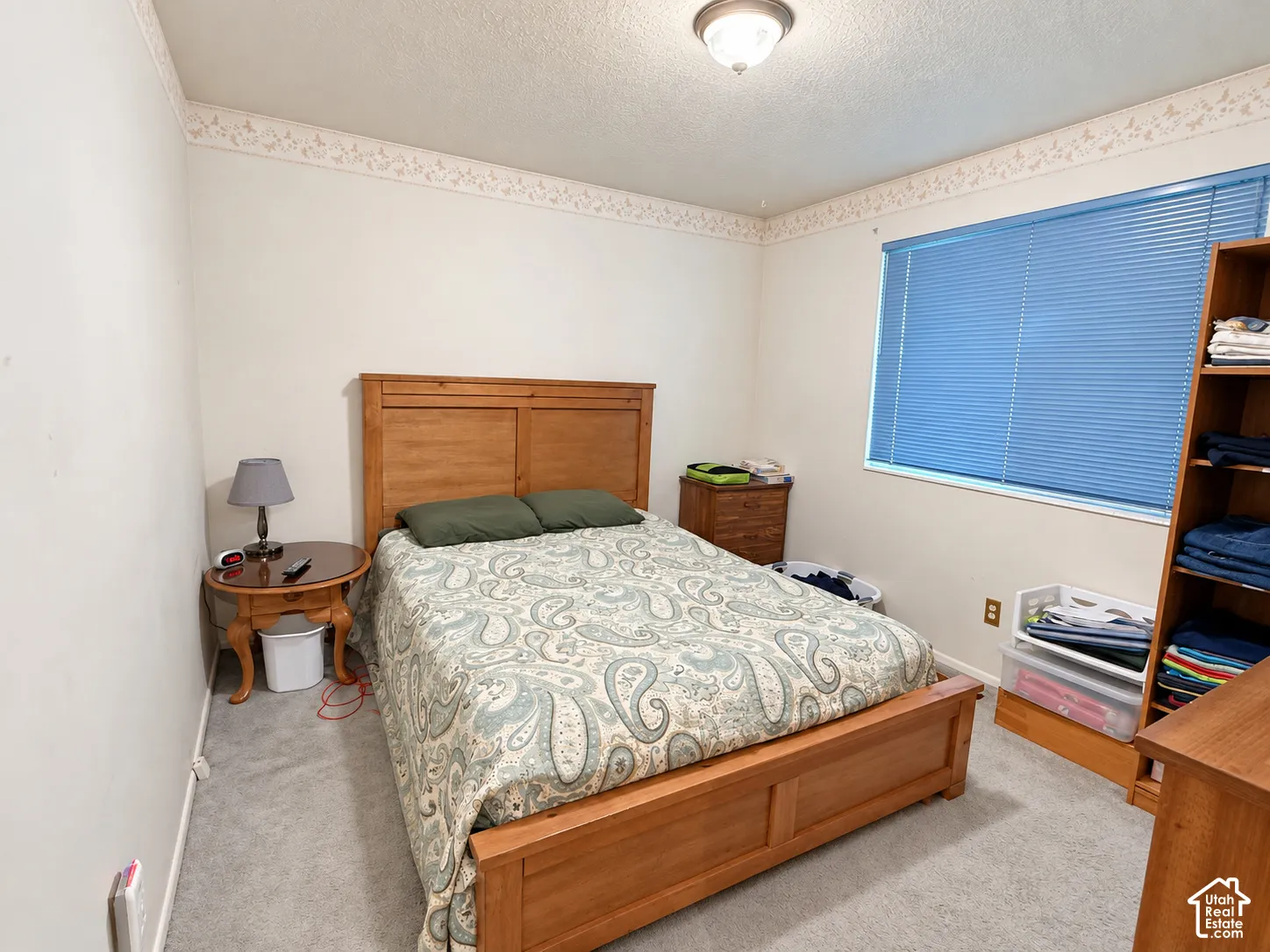 Spacious bedroom featuring warm wood-finish furnishings, carpeted flooring, and abundant natural light from a large window with blue blinds