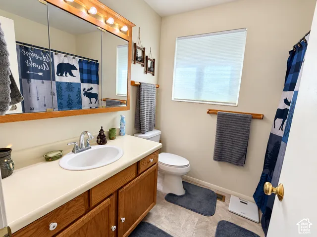 Bathroom featuring a single-sink vanity with light wood cabinetry, a large mirror with integrated lighting, a commode, and a window with blinds