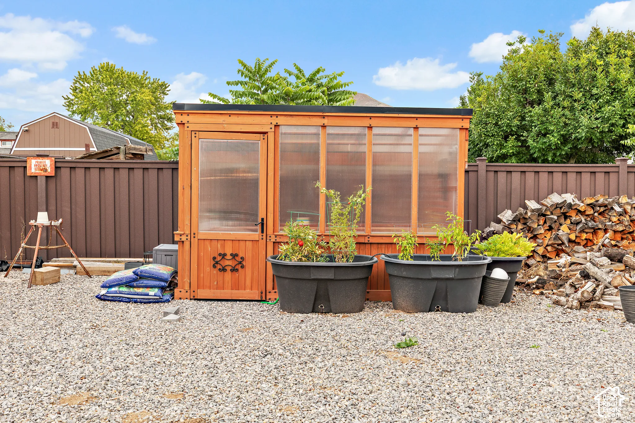 Freestanding wooden greenhouse with translucent panels, set on a gravel surface, alongside a privacy fence