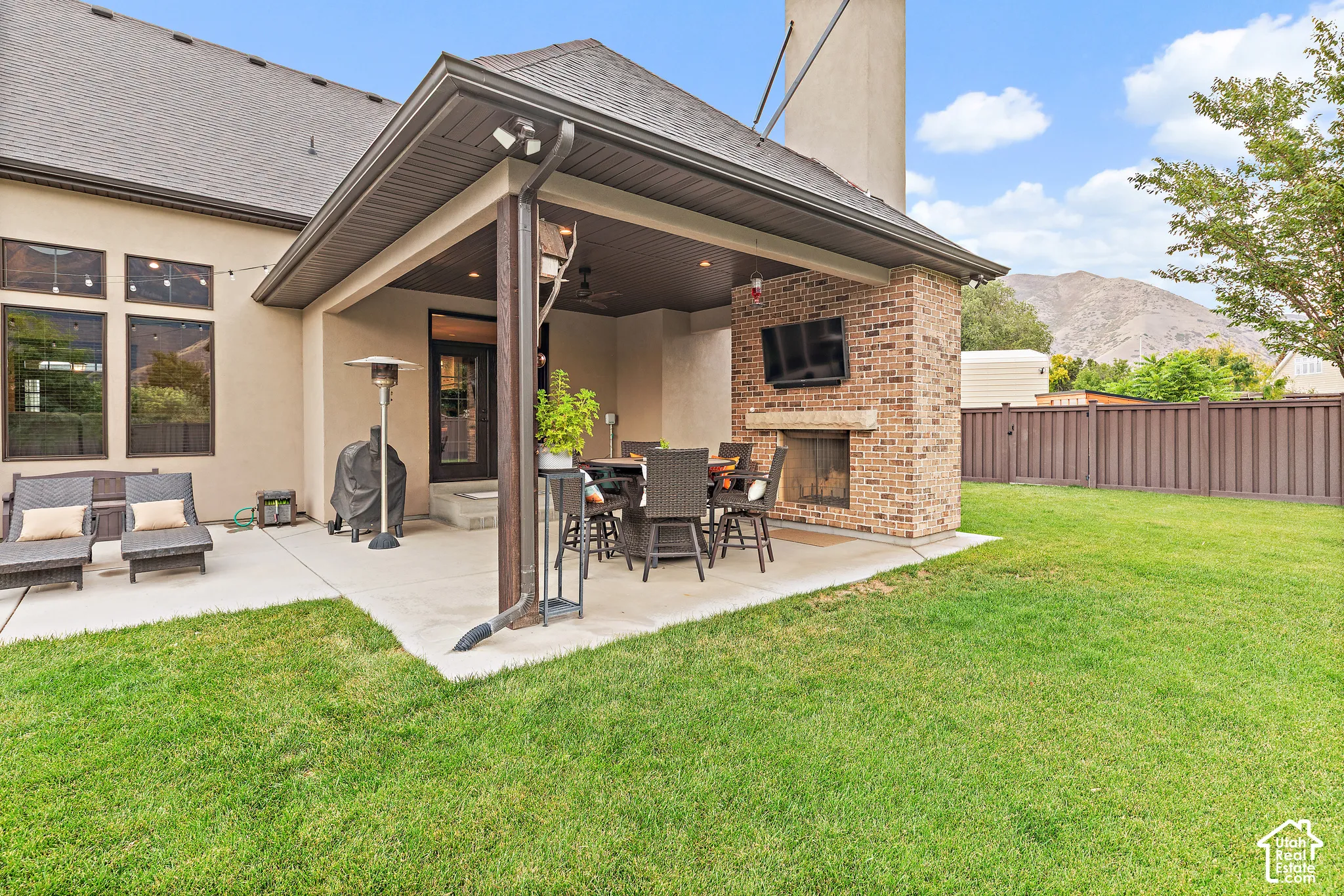 Expansive concrete patio featuring a covered outdoor living area with a brick fireplace, built-in outdoor television, and mountain backdrop
