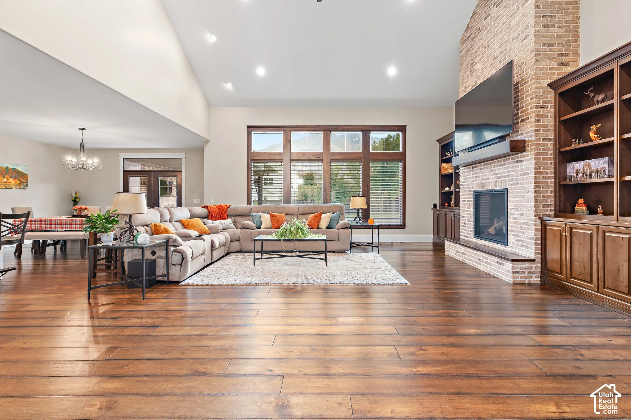 High vaulted ceiling, wood-finish flooring, brick-clad fireplace, built-in shelving, and large multi-pane windows