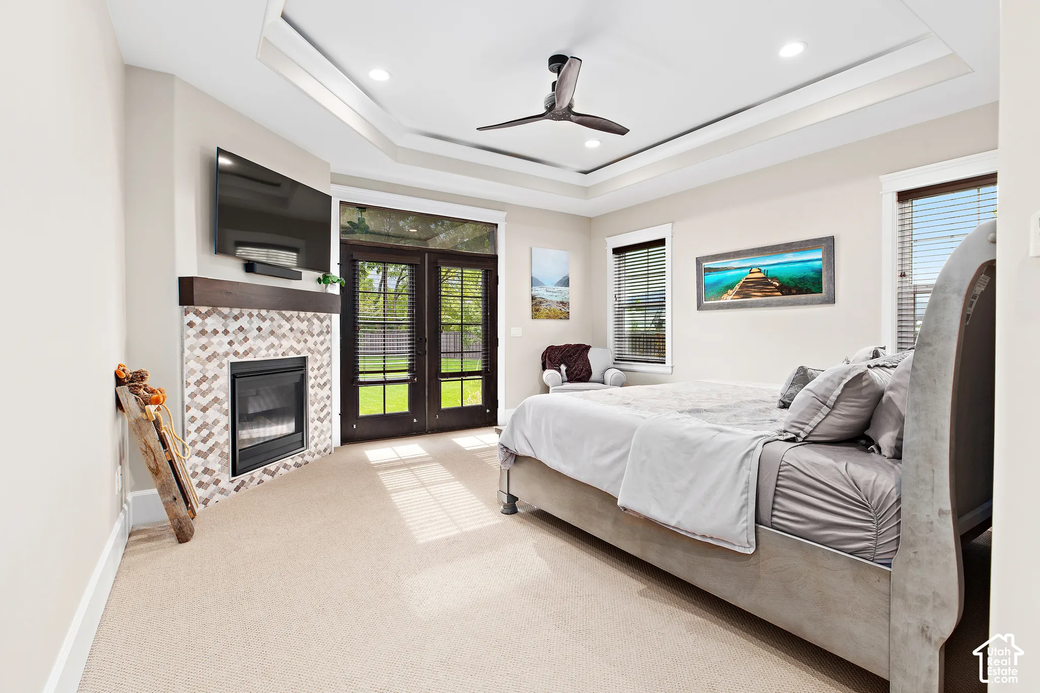Tray ceiling with recessed lighting and a ceiling fan, corner fireplace with a decorative tile surround and wood mantel, light-colored carpeting, and French doors