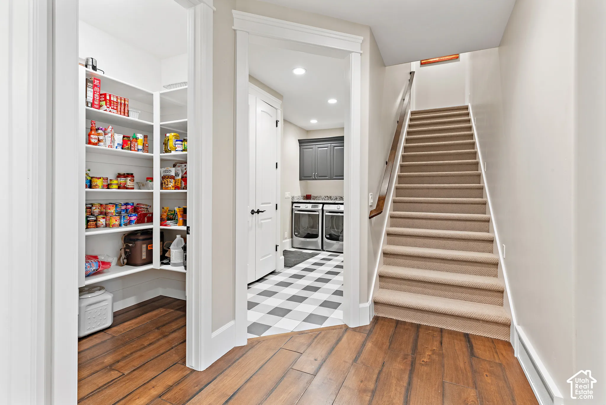 Walk-in pantry with extensive built-in shelving, wood-finish flooring, carpeted staircase, and tiled floor entry to a utility area