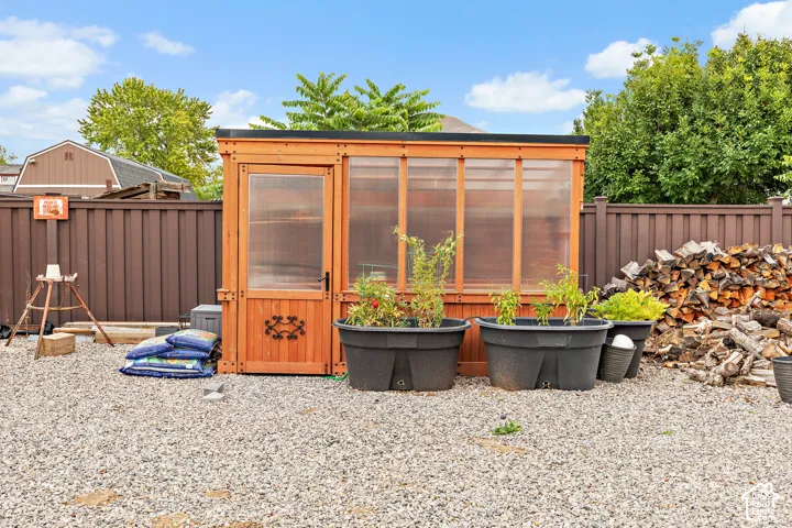 Freestanding wooden greenhouse with translucent panels, set on a gravel surface, alongside a privacy fence