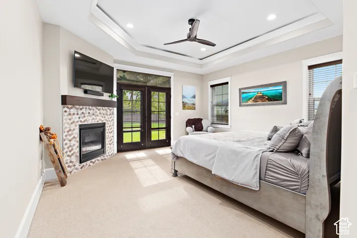 Tray ceiling with recessed lighting and a ceiling fan, corner fireplace with a decorative tile surround and wood mantel, light-colored carpeting, and French doors