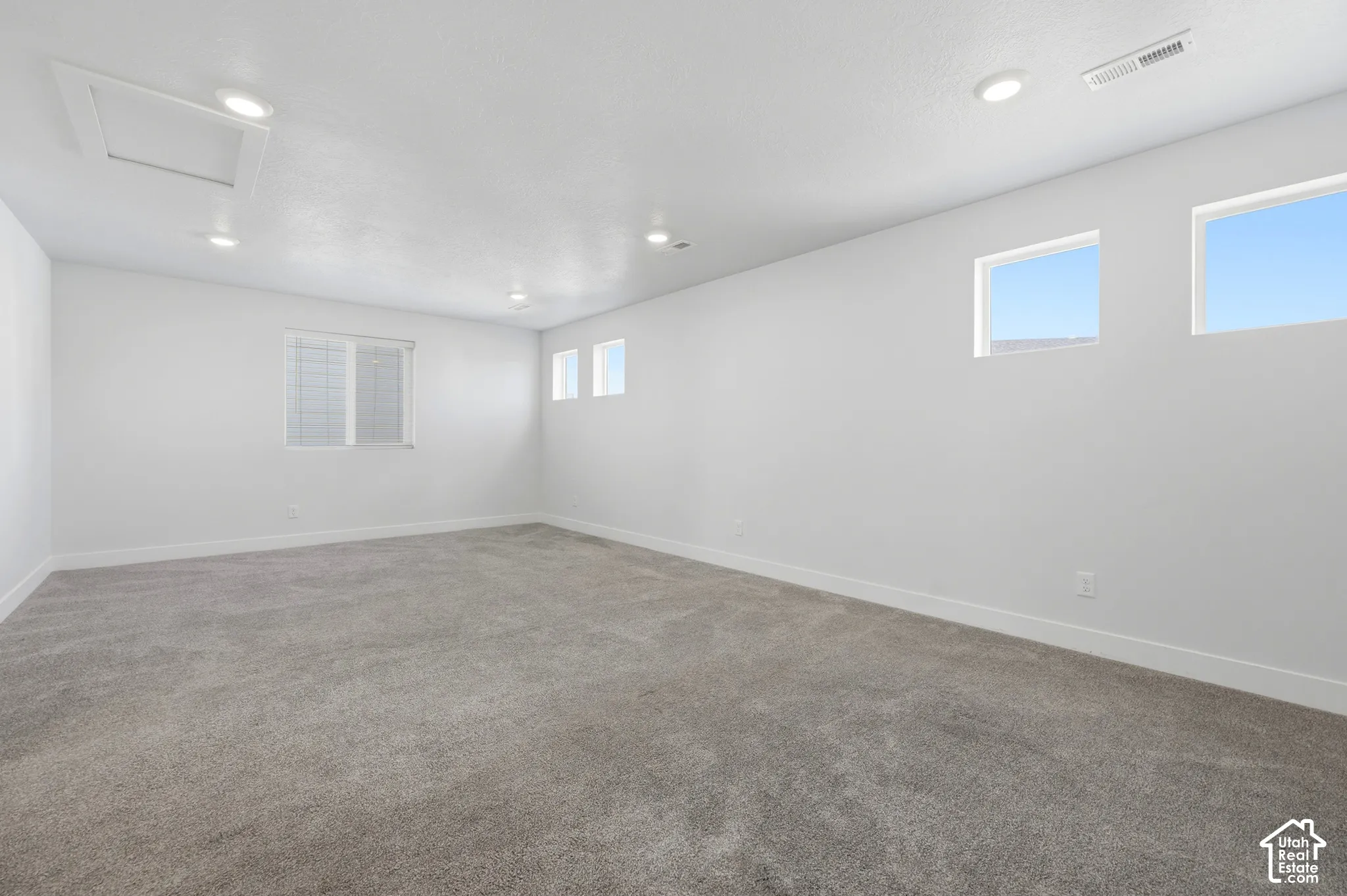 Expansive room featuring plush gray carpeting, recessed ceiling lighting, and multiple windows providing natural illumination