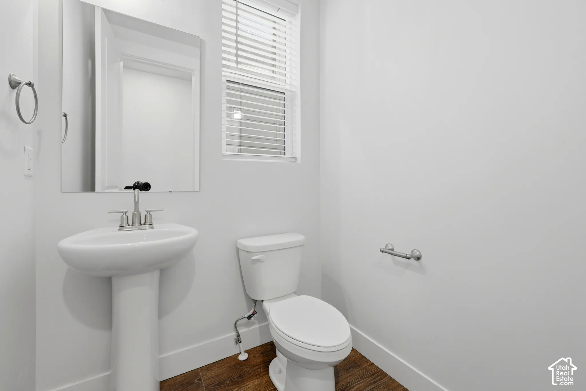 Pedestal sink, wall-mounted mirror, wood-finish flooring, window with blinds, and white toilet