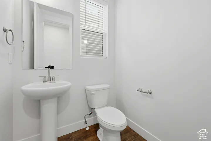 Pedestal sink, wall-mounted mirror, wood-finish flooring, window with blinds, and white toilet