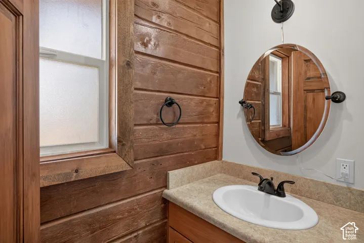 Wood-paneled wall, single-pane window with privacy glass, vanity with an integrated sink, dual-handle faucet, and round wall mirror