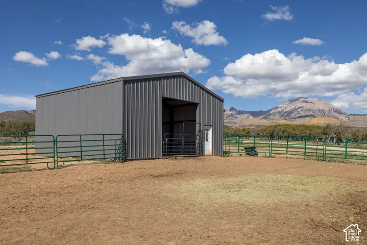 Modern metal-frame agricultural structure featuring a spacious open-front design with sliding barn doors, sturdy pipe fencing, and expansive dirt work area
