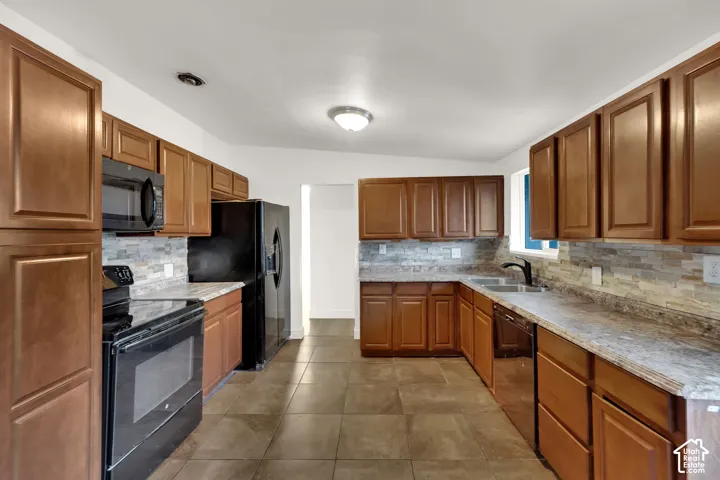 Kitchen featuring extensive wood cabinetry, tile flooring, black appliances, stone-look countertops, and a tile backsplash