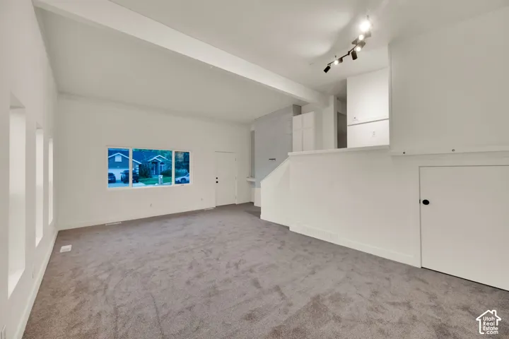 Vaulted ceiling with exposed beam, multiple windows, track lighting, and soft gray carpeting