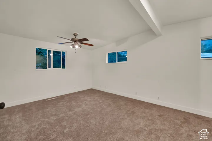 Expansive room featuring plush carpeting, multiple windows, a ceiling fan, and a prominent ceiling beam