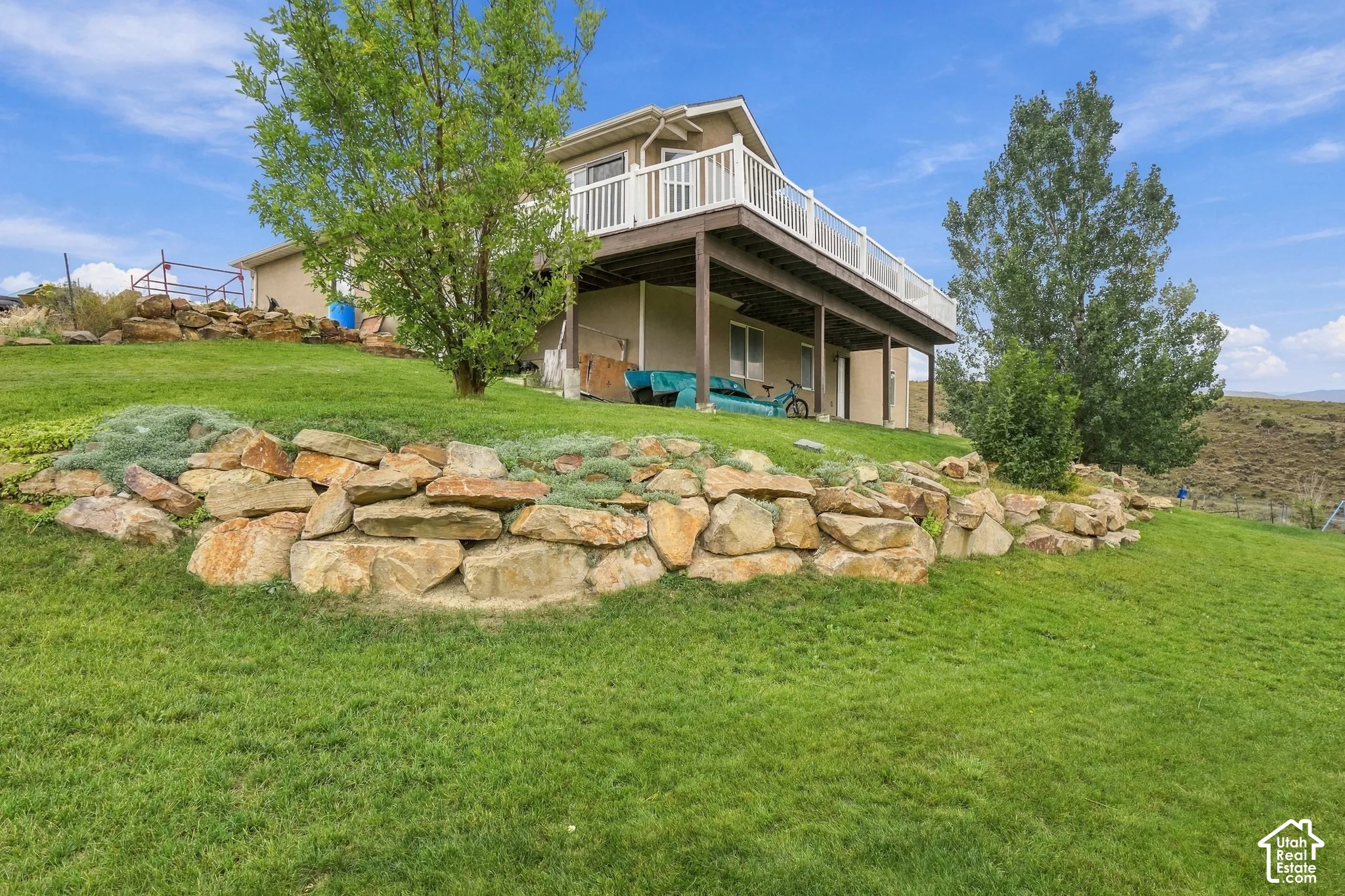 Expansive green lawn, multi-level rock retaining wall, mature trees, upper-level deck with white railing, and lower-level covered patio