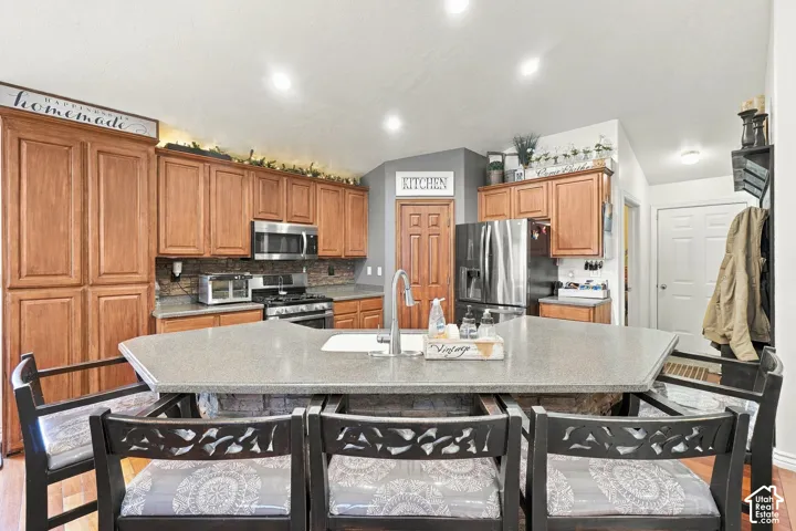 Kitchen featuring a central island with an integrated sink, wood-finish cabinetry, stainless steel appliances, recessed lighting, and a stone-finish backsplash