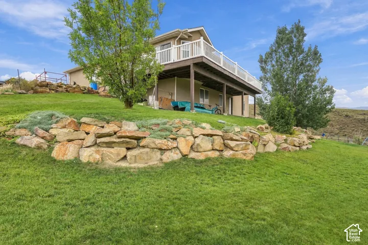 Expansive green lawn, multi-level rock retaining wall, mature trees, upper-level deck with white railing, and lower-level covered patio