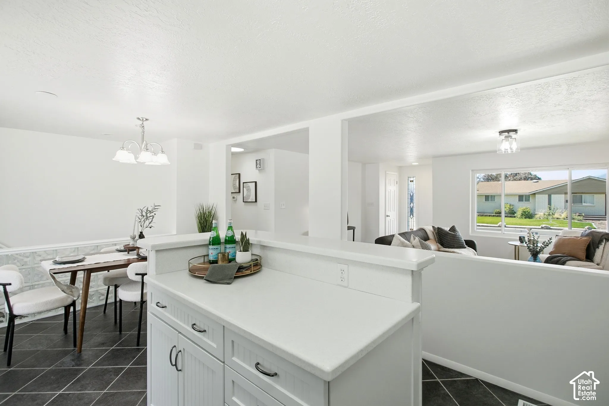 White kitchen island with solid surface countertop, dark tile flooring, dining area with chandelier, and a living area with a large window