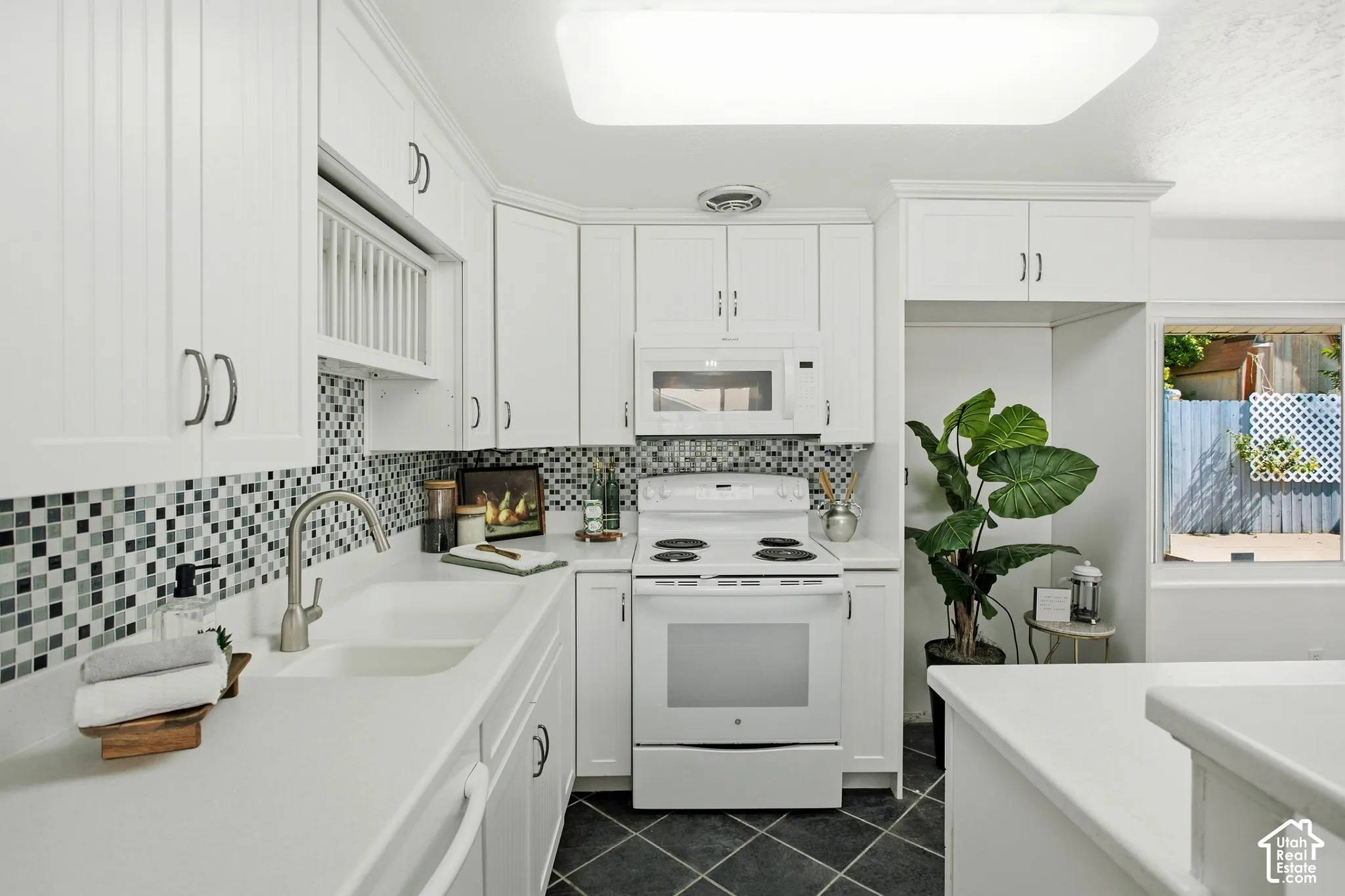 White cabinetry with dark hardware, mosaic tile backsplash, double basin sink with a gooseneck faucet, white built-in appliances, and dark diamond-patterned flooring